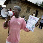 Matthew Zawalo hält in einer Hand ein Megaphon, in der anderen ein Plakat. Er arbeitet als Community Health Worker in Liberia. Auf dem Markt des Dorfes Busie wirbt er für eine Infoveranstaltung über das Corona-Virus. Foto: Christoph Pueschner/Brot für die Welt
