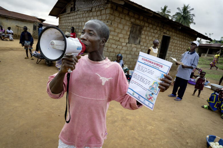 Matthew Zawalo hält in einer Hand ein Megaphon, in der anderen ein Plakat. Er arbeitet als Community Health Worker in Liberia. Auf dem Markt des Dorfes Busie wirbt er für eine Infoveranstaltung über das Corona-Virus. Foto: Christoph Pueschner/Brot für die Welt
